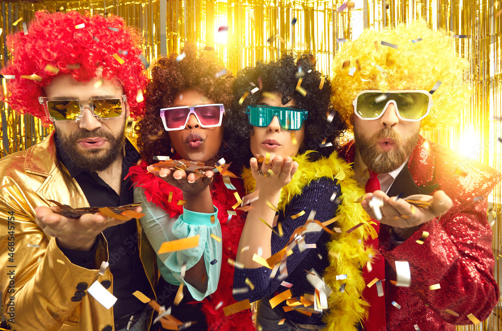 Happy merry diverse people dressed in boas, glasses and curly Afro wigs celebrating New Year at disco night club party throw a kiss blowing shiny golden confetti sharing groovy festive mood with you