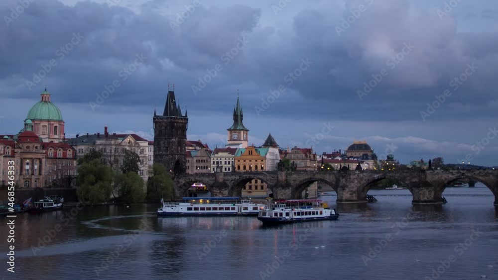 Day to night sunset timelapse with a close up view of Charles Bridge on Vltava river with boats on a cloudy evening in Prague, Czech Republic as the bridge and towers of the city light up