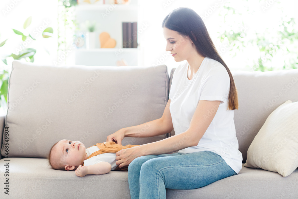 Photo of pretty charming lady wear white t-shirt sitting couch smiling dressing baby inside indoors house room Photo of pretty charming lady wear white t-shirt sitting couch smiling dressing baby inside indoors house room