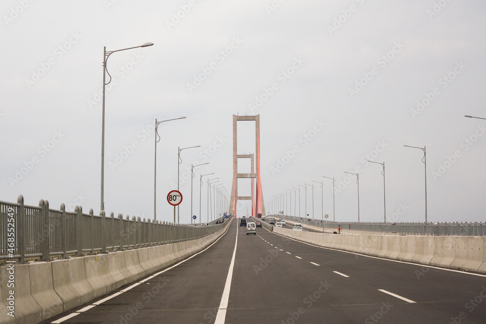 Scene of the famous Suramadu Bridge and its red suspension steel cables with cars and lamp post on road and cloudy sky background. 