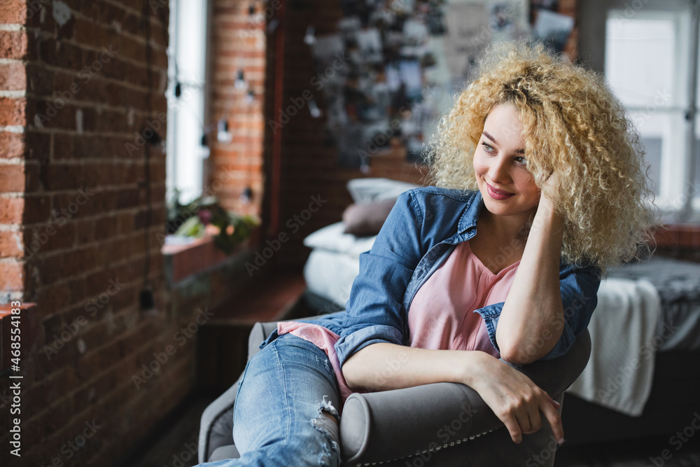 Cute charming curly blonde in modern apartment in a cozy armchair looks to the side Cute charming curly blonde in modern apartment in a cozy armchair looks to the side