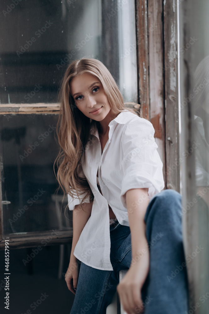 a beautiful girl in a white shirt with blonde hair is sitting on the windowsill of a large window a beautiful girl in a white shirt with blonde hair is sitting on the windowsill of a large window