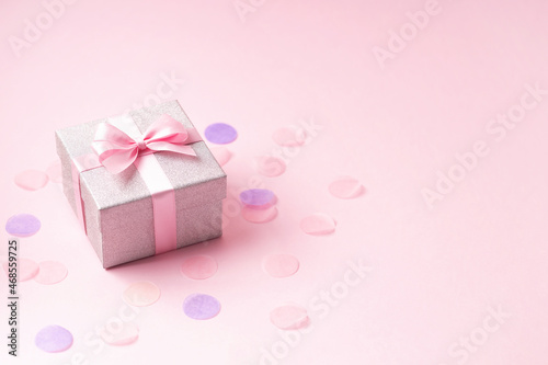 Silver glitter gift box with pink ribbon bow on pink background with confetti.