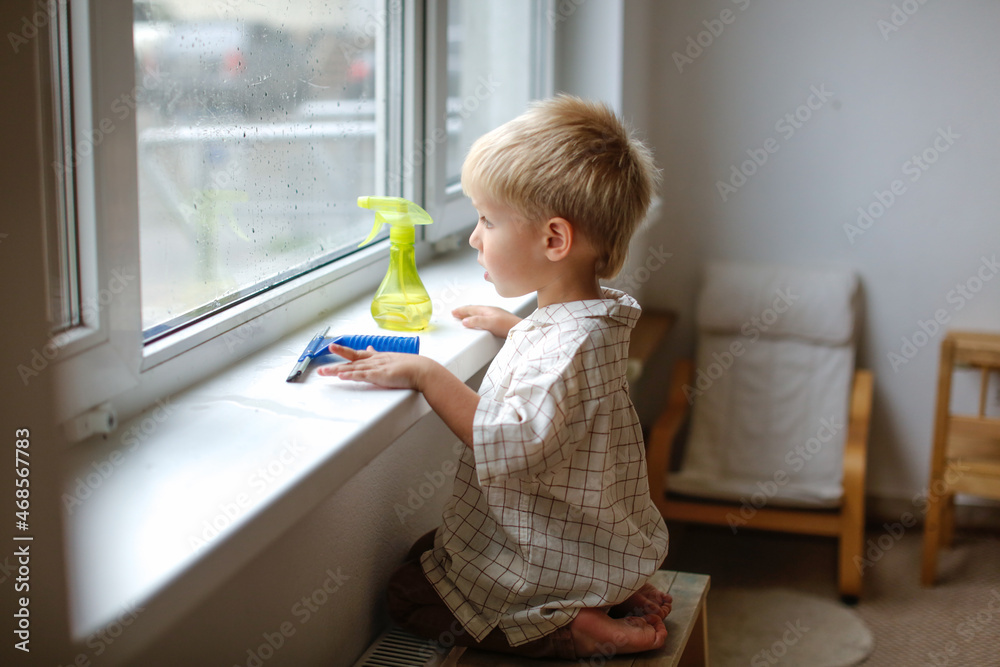 Cute boy toddler blond toddler at the window washes the glass. Helping children with household chores and household chores, children's independence, Montessori classes for small Cute boy toddler blond toddler at the window washes the glass. Helping children with household chores and household chores, children's independence, Montessori classes for small