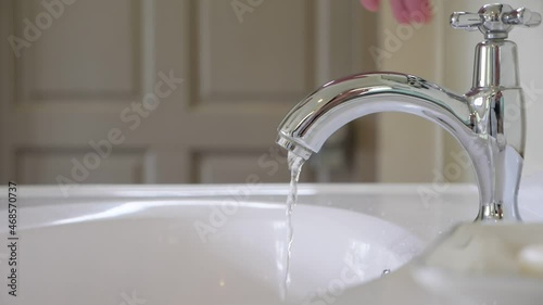 A thin trickle of water pours from the faucet in sink at bathroom, man closes the tap and stop the leakage. Close up slow motion shot of polished spigot, blurred door on background