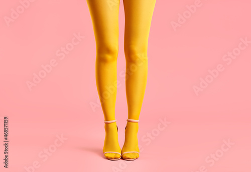 Long slender legs of young woman in yellow tights and stylish high heels shoes on pink background.