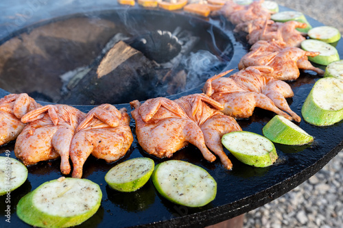 Close-up detail view of many tasty raw marinated quails and vegetables and mushrooms grilled cooked at round steel iron firepit hearth table surface brazier with burning firewoods. Barbecue yard home photo