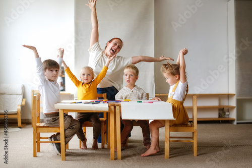 cheerful teacher woman with a group of cute preschool children at the table draw on paper with multicolored felt-tip pens, montessori and creativity, creative development and lesson in kindergarten