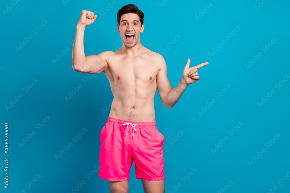 Photo of excited guy direct finger empty space rejoice win wear swimwear shorts isolated blue color background