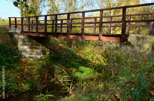 pedestrian bridge over a small gorge and a stream. it consists of two steel crossbeams. as the surface and railing of the bridge is made of brown painted planks. is long and narrow. in the wild