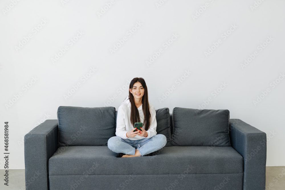 Smiling young woman using phone, sitting on couch at home, looking at smartphone screen, beautiful female chatting in social network or shopping online, spending leisure time with mobile device Smiling young woman using phone, sitting on couch at home, looking at smartphone screen, beautiful female chatting in social network or shopping online, spending leisure time with mobile device
