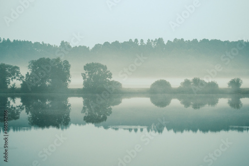 misty morning by the lake with calm water, fog and reflections of trees in mirror