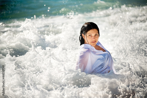 A girl with wet hair in a white shirt in the foam of the waves against the background of the azure waves of the sea. 