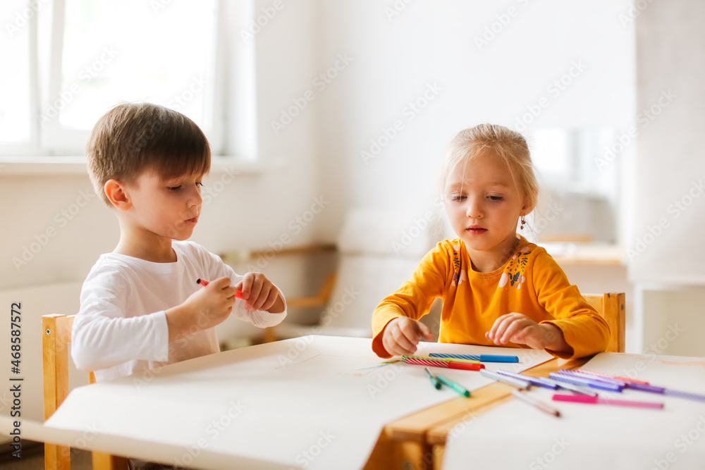 Funny kids Toddlers draw together with felt-tip pens on paper at the table, Montessori and creativity, creative development and activities in kindergarten. Boy and girl painting together Funny kids Toddlers draw together with felt-tip pens on paper at the table, Montessori and creativity, creative development and activities in kindergarten. Boy and girl painting together