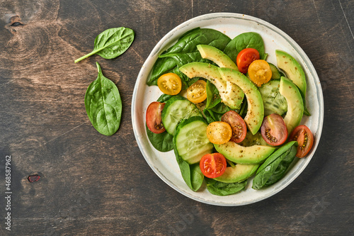 Avocado, red, yellow, black cherry tomato, spinach and cucumber fresh salad with spices pepper and olive oil in grey bowl old wooden table background. Healthy food concept. Top view.
