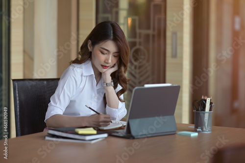 Free Download Photo of a young businesswoman keeping a hand on her chin while taking notes at the wooden meeting table surrounded by a digital tablet and office equipment in the comfortable meeting room. - GFXLoad
