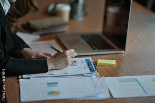 Close-up image of a young businesswoman writing on a paperwork at the wooden table surrounded by paperwork, office equipment and computer laptop.