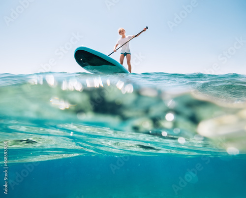 Under the water view angle to the smiling blonde teenager, boy rowing stand up paddleboard. Active family summer vacation time near the sea concept image. photo