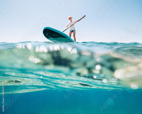 Fotografie Under the water view angle to the smiling blonde teenager, boy rowing stand up paddleboard