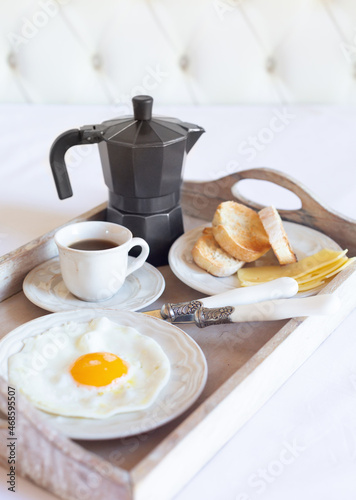 A breakfast tray with a fried egg, a cup of black coffee and a coffee pot is on a white bed. Vertical image.