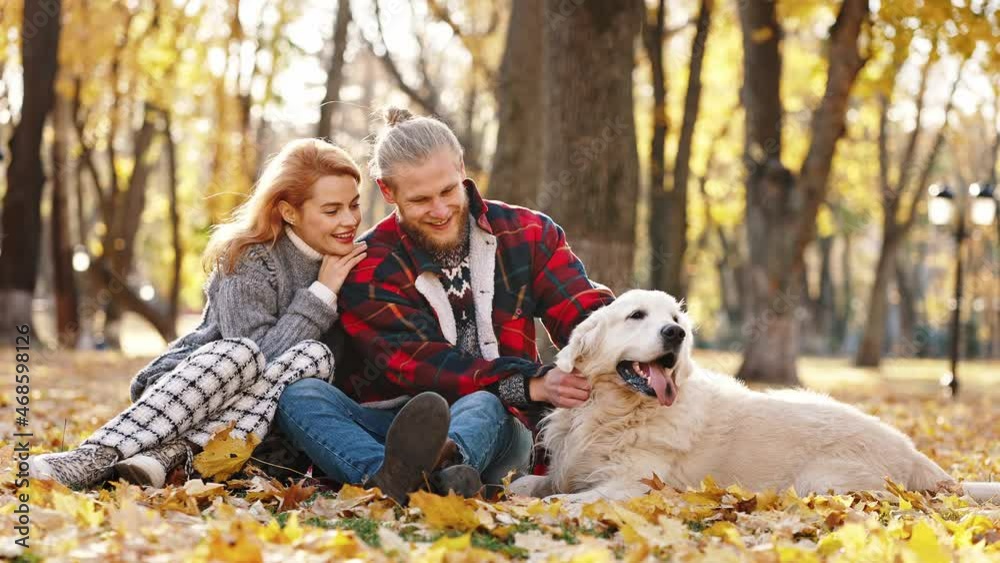 Family and love. Young married man and woman caressing dog, resting on fallen leaves in autumn park, slow motion Family and love. Young married man and woman caressing dog, resting on fallen leaves in autumn park, slow motion