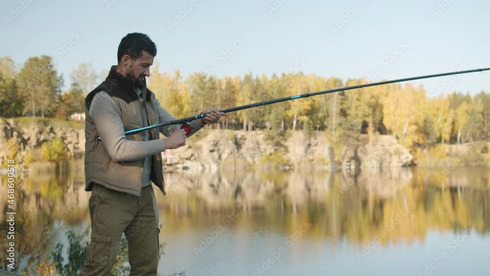 Young fisherman in casual clothing is catching fish with rod standing against natural background alone watching beautiful landscape. People and hobby concept. Young fisherman in casual clothing is catching fish with rod standing against natural background alone watching beautiful landscape. People and hobby concept.