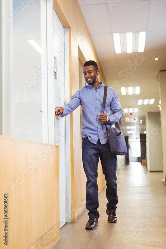 Smiling african american businessman walking and opening door in corridor in modern office
