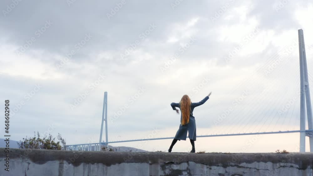 Aerial panoramic view of a woman with long hair dancing in artistic manner. The Russian bridge across Eastern Bosphorus strait, Vladivostok cityscape, coastline and sea port with vessels on background Aerial panoramic view of a woman with long hair dancing in artistic manner. The Russian bridge across Eastern Bosphorus strait, Vladivostok cityscape, coastline and sea port with vessels on background