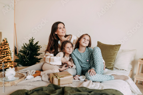 A happy mom with two daughters is resting, relaxing and having fun sitting on the bed in a cozy decorated bedroom during the Christmas holiday at home during the New Year vacations. Selective focus © Елизавета Старкова