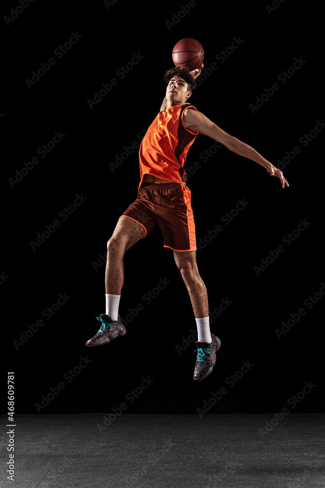 Full length portrait of basketball player training isolated on dark studio background. Tall muscular athlete jumping with ball.