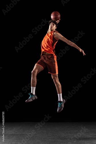 Full length portrait of basketball player training isolated on dark studio background. Tall muscular athlete jumping with ball. © master1305