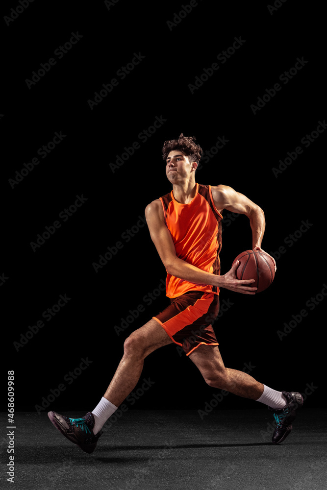 Full length portrait of basketball player training isolated on dark studio background. Tall muscular athlete jumping with ball.