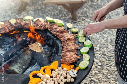 Close-up detail view of chef flipping by tongs tasty crispy marinated quails and vegetables grilled at round steel iron firepit hearth table surface brazier with burning firewoods. Barbecue yard home