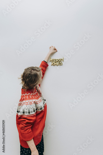 a modern woman in a red sweater hangs a Christmas inscription on a white wall. MERRY XMAS. The minimum concept of the new year