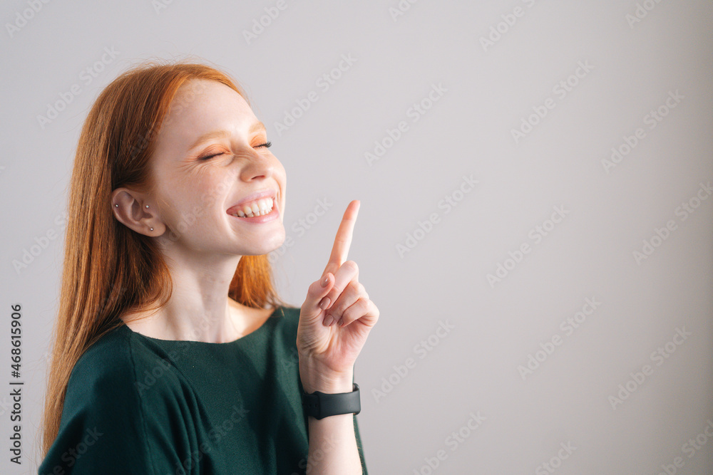 Portrait of laughing redhead young woman model pointing up with finger with closed eyes on white background. Medium shot of red-haired lady with natural beauty posing in professional studio. Portrait of laughing redhead young woman model pointing up with finger with closed eyes on white background. Medium shot of red-haired lady with natural beauty posing in professional studio.