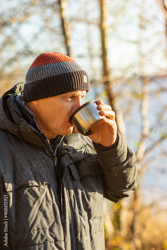 An elderly man drinks hot tea from a thermos on the shore of a lake with colorful autumn nature in the background. Hot coffee drink in nature.