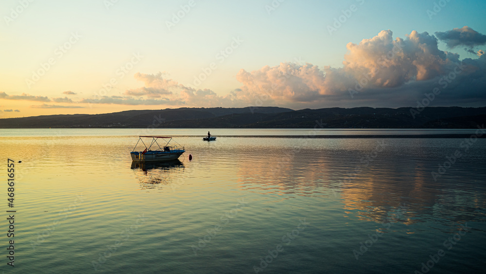 Couché de soleil sur un petit bateau sur un lac, les nuages se reflètent sur l'eau, de couleur orange et bleu. Une colline en face. Couché de soleil sur un petit bateau sur un lac, les nuages se reflètent sur l'eau, de couleur orange et bleu. Une colline en face.