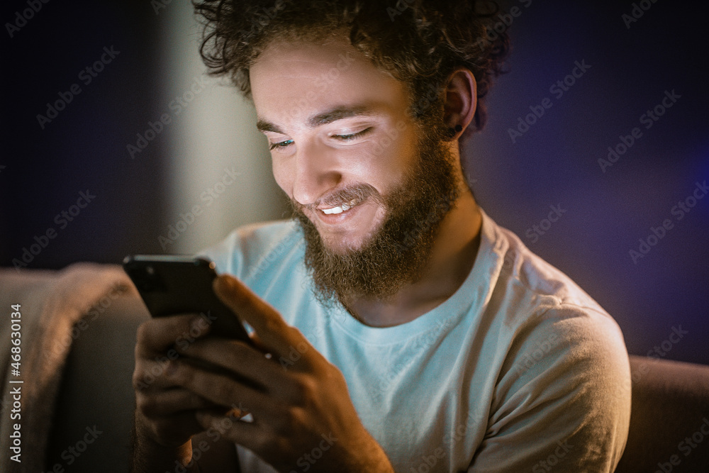 Curly Guy Swipes on His Phone Sitting on the Couch. Nice Man is Typing a Message to His Girlfriend on His Phone. Close up portrait Curly Guy Swipes on His Phone Sitting on the Couch. Nice Man is Typing a Message to His Girlfriend on His Phone. Close up portrait