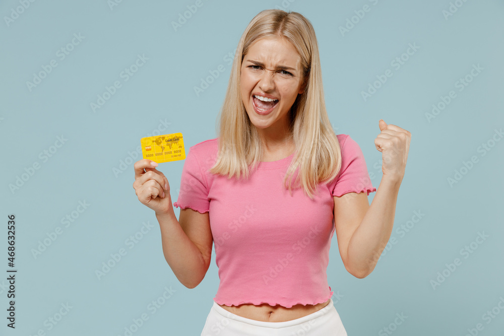 Young excited smiling happy cheerful caucasian blonde woman 20s in casual pink t-shirt hold in hand credit bank card do winner gesture isolated on plain pastel light blue background studio portrait. Young excited smiling happy cheerful caucasian blonde woman 20s in casual pink t-shirt hold in hand credit bank card do winner gesture isolated on plain pastel light blue background studio portrait.