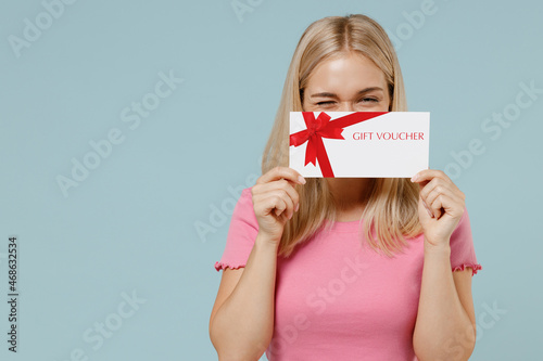 Full body fun young blonde woman 20s wearing casual pink t-shirt sit in bag chair cover mouth with gift certificate coupon voucher card for store isolated on plain pastel light blue background studio.