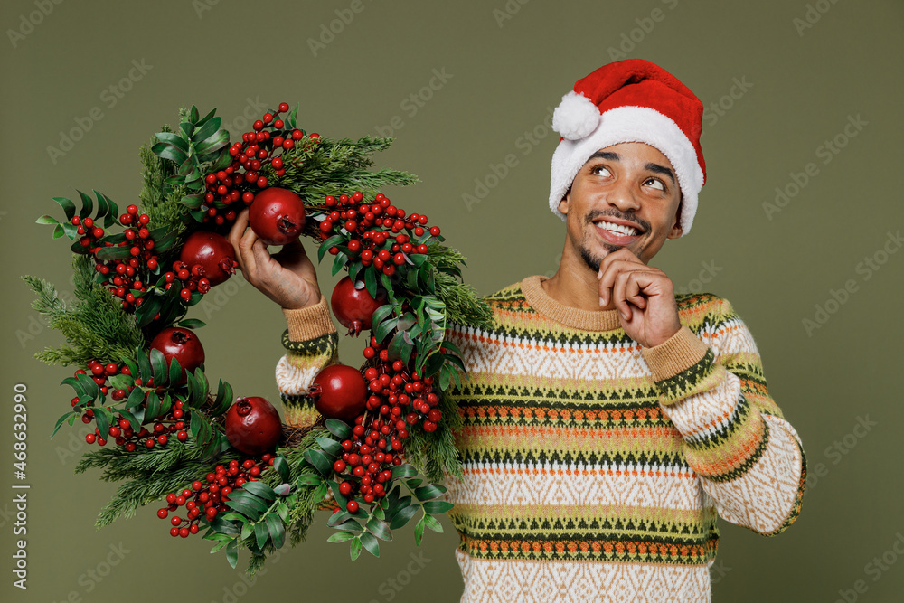 Young minded fun african man in knitted sweater red Christmas hat hold Advent wreath look overhead prop up chin isolated on plain green khaki background studio Happy New Year 2022 celebration concept.