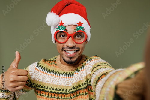Young close up african man in sweater red Christmas hat fun glasses do selfie shot pov on mobile phone show thumb up isolated on plain green khaki background. Happy New Year 2022 celebration concept.