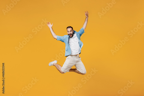 Full body young smiling happy caucasian man 20s wearing blue shirt white t-shirt jump high with outstretched hands arms isolated on plain yellow background studio portrait. People lifestyle concept.