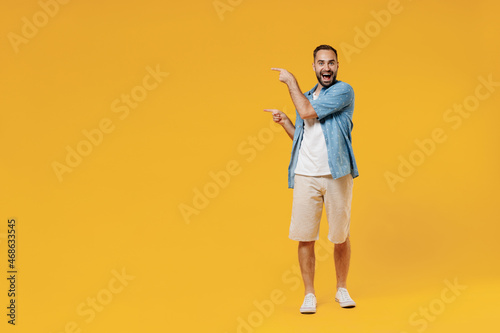 Full body young smiling fun happy caucasian man 20s wearing blue shirt white t-shirt point index finger aside on workspace area mock up copy space isolated on plain yellow background studio portrait