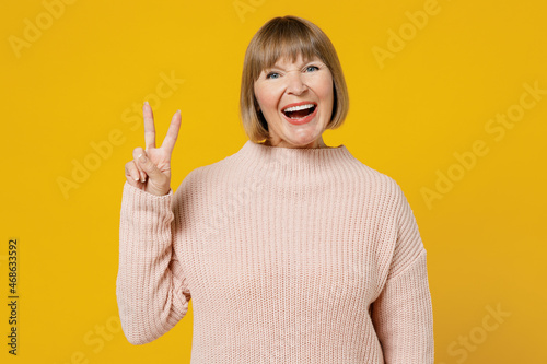 Elderly smiling happy fun caucasain cheerful woman 50s wears pink casual knitted sweater showing victory sign look camera isolated on plain yellow background studio portrait. People lifestyle concept. photo