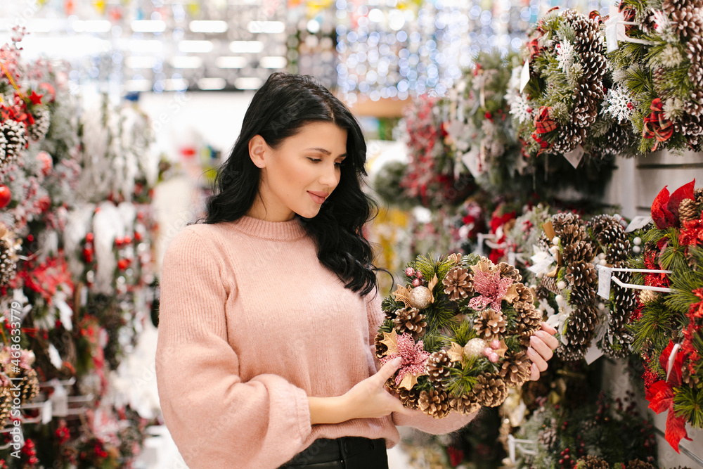Young brunette woman in a pink sweater chooses a Christmas wreath in the store. New Year's shopping.