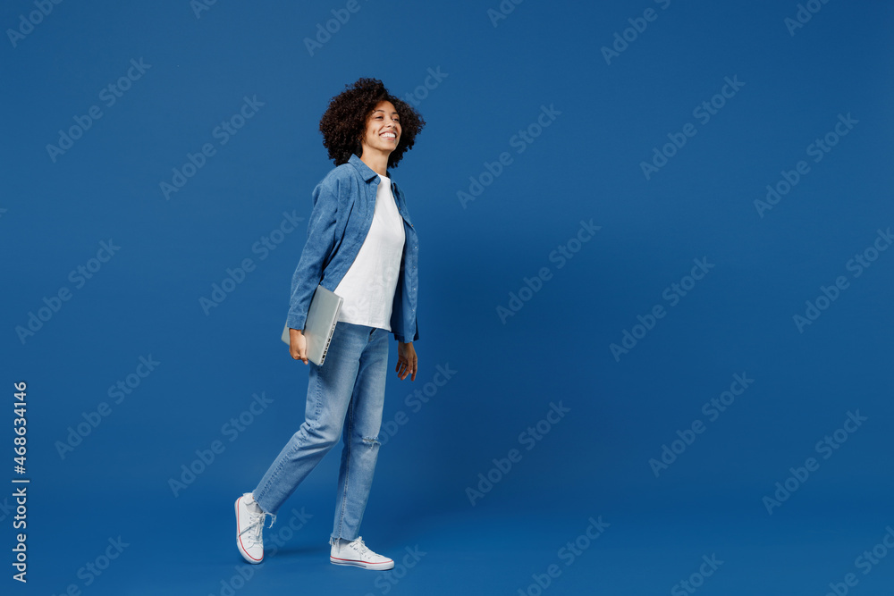 Full body side view fun young smiling happy black woman in casual clothes shirt white t-shirt hold closed laptop pc computer isolated on plain dark blue background studio. People lifestyle concept