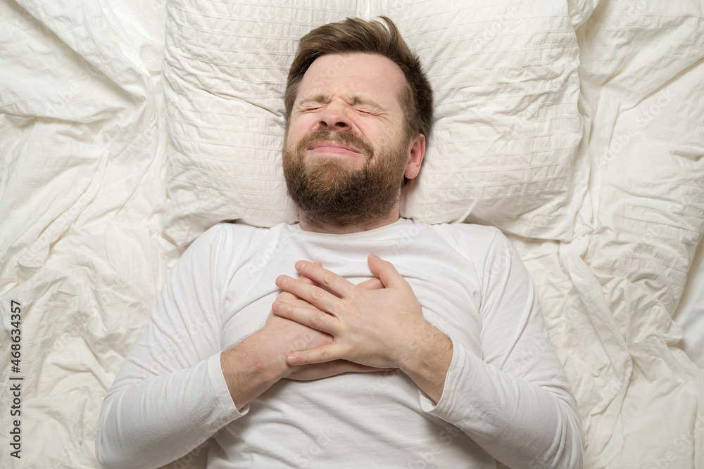 Man in white pajamas is suffering from severe heartache, he is suffering and has hands on his chest. Top view. Close-up. Man in white pajamas is suffering from severe heartache, he is suffering and has hands on his chest. Top view. Close-up.