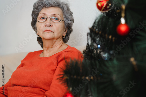 Portrait of lonely sad senior woman with glasses sitting alone at home near xmas tree and looking away. Loneliness in Christmas celebration, self-isolation, lockdown for holiday