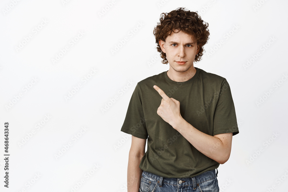 Angry curly guy pointing left, frowning and looking displeased, complaining at smth, standing in green t-shirt over white background Angry curly guy pointing left, frowning and looking displeased, complaining at smth, standing in green t-shirt over white background